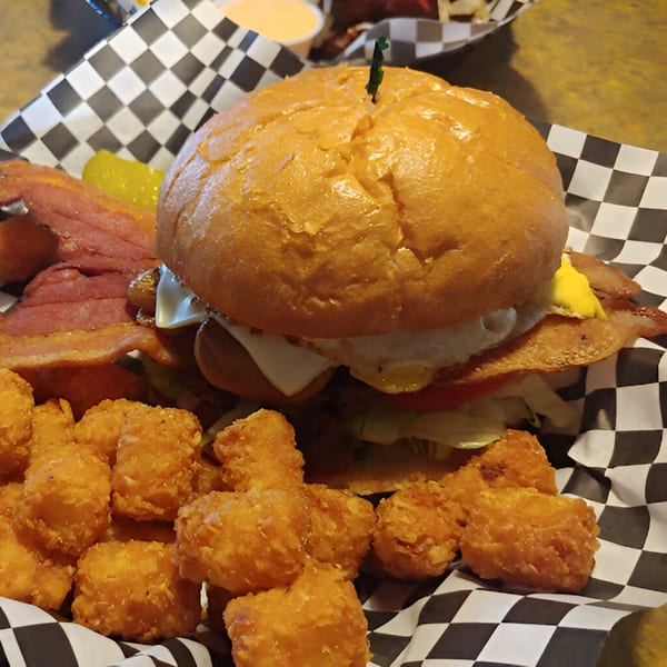 A large burger with bacon, egg, cheese, and vegetables, served with tater tots in a checkered basket.